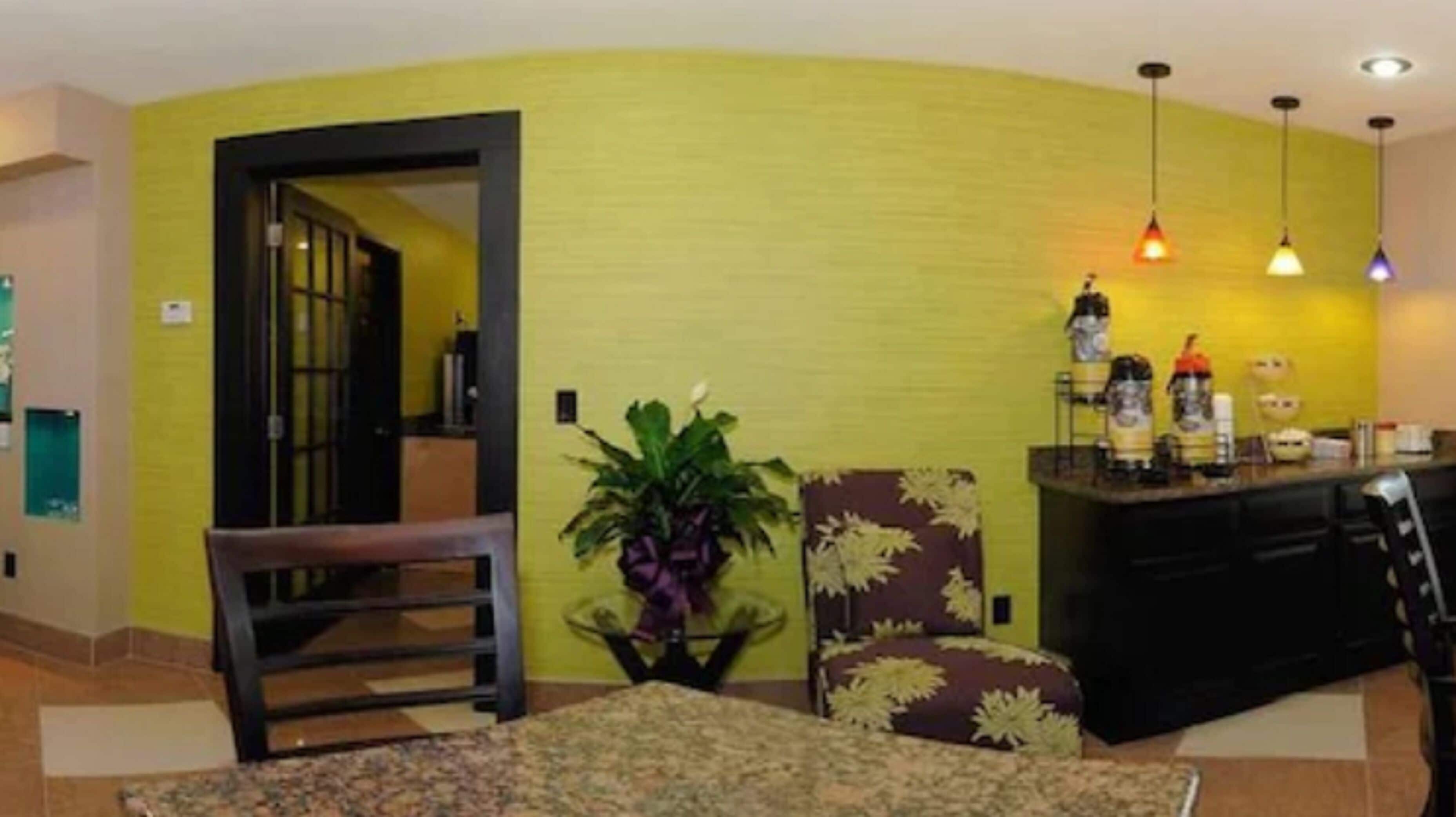 Una sala de estar con pared amarilla, un comedor con mesa y sillas negras, y una cocina con mostrador y luces colgantes.