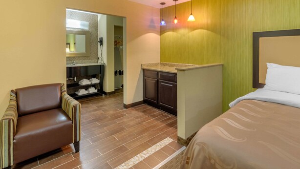 A hotel room with a bed, a chair, a small kitchenette, and a bathroom visible through an open door.