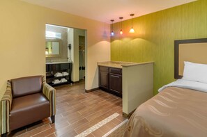 A hotel room with a bed, a chair, a small kitchenette, and a bathroom visible through an open door.