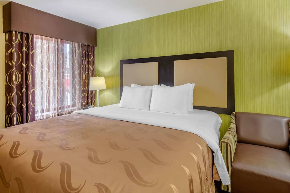 Quarto de hotel com uma cama grande, cabeceira, mesa de cabeceira, luminária e uma poltrona de couro marrom.