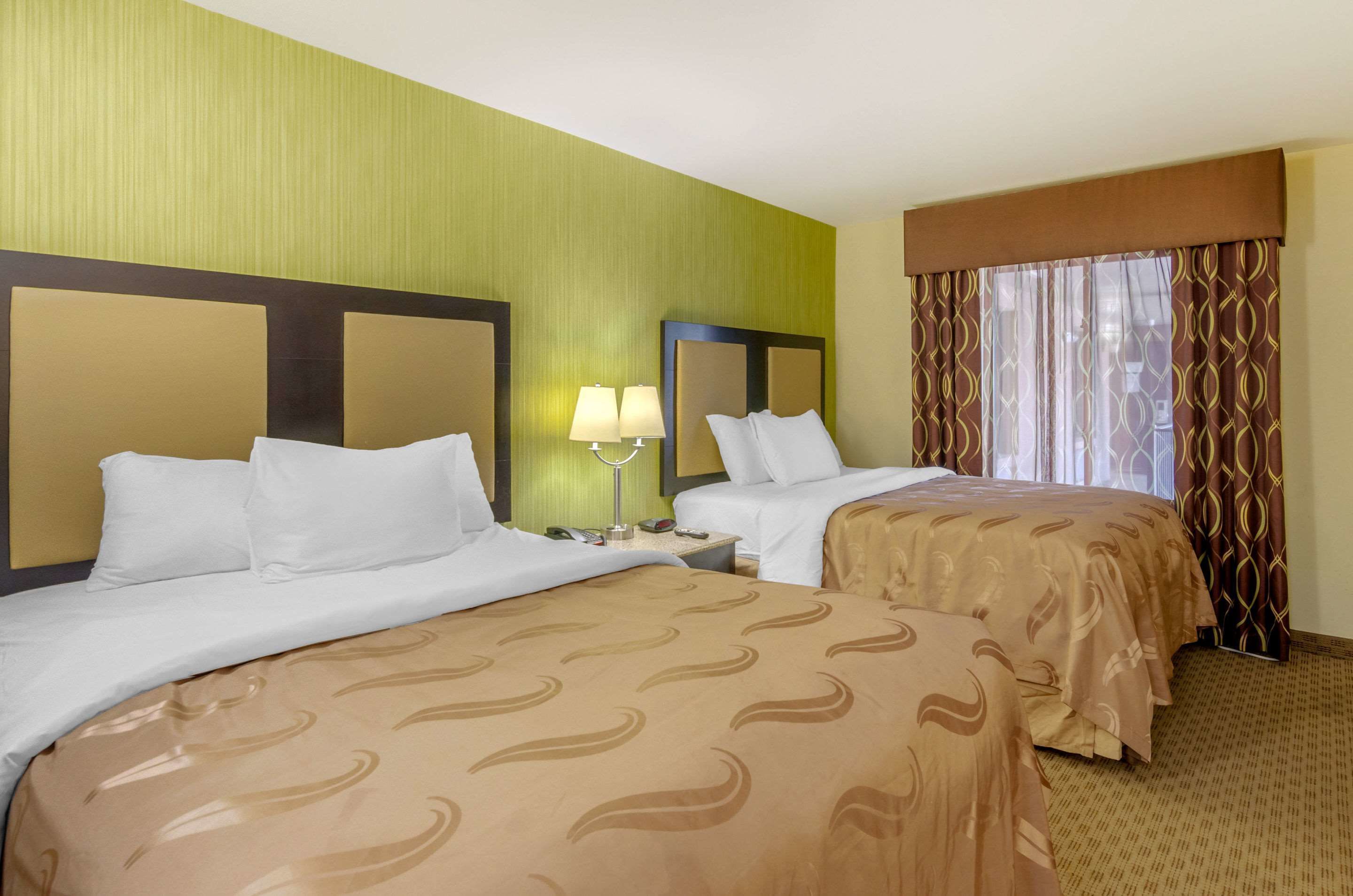 A hotel room with two beds, a nightstand with a lamp, and a window with patterned curtains.