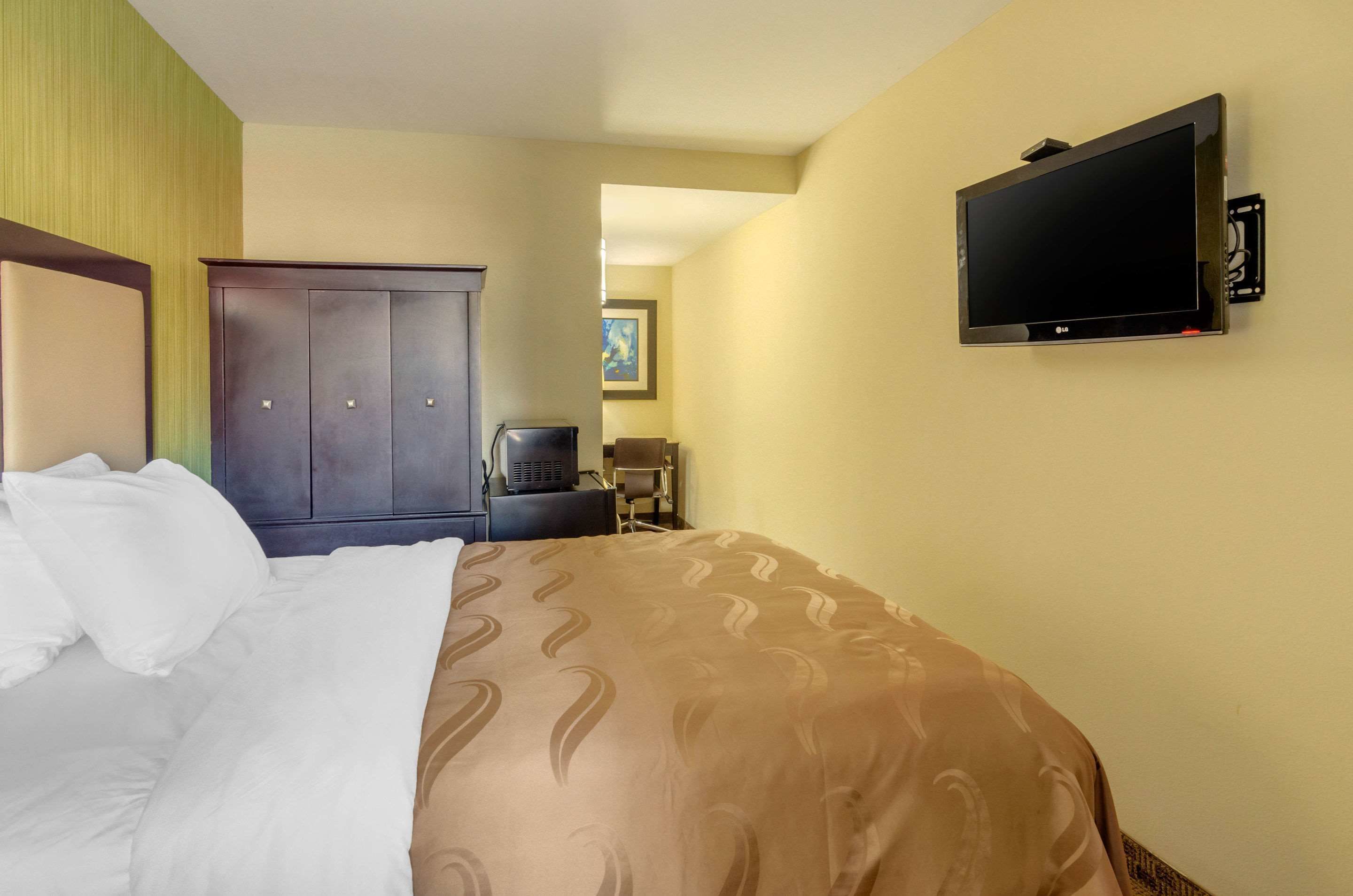 Una habitación de hotel con cama, armario, televisor montado en la pared y una pequeña mesa con silla.