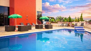 A swimming pool with lounge chairs and umbrellas, surrounded by a red building and palm trees.