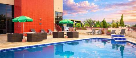 A swimming pool with lounge chairs and umbrellas, surrounded by a red building and palm trees.