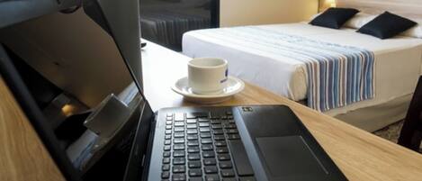 A laptop on a wooden desk with a cup on a saucer, a bed with a striped blanket, and a window with curtains in a hotel room.