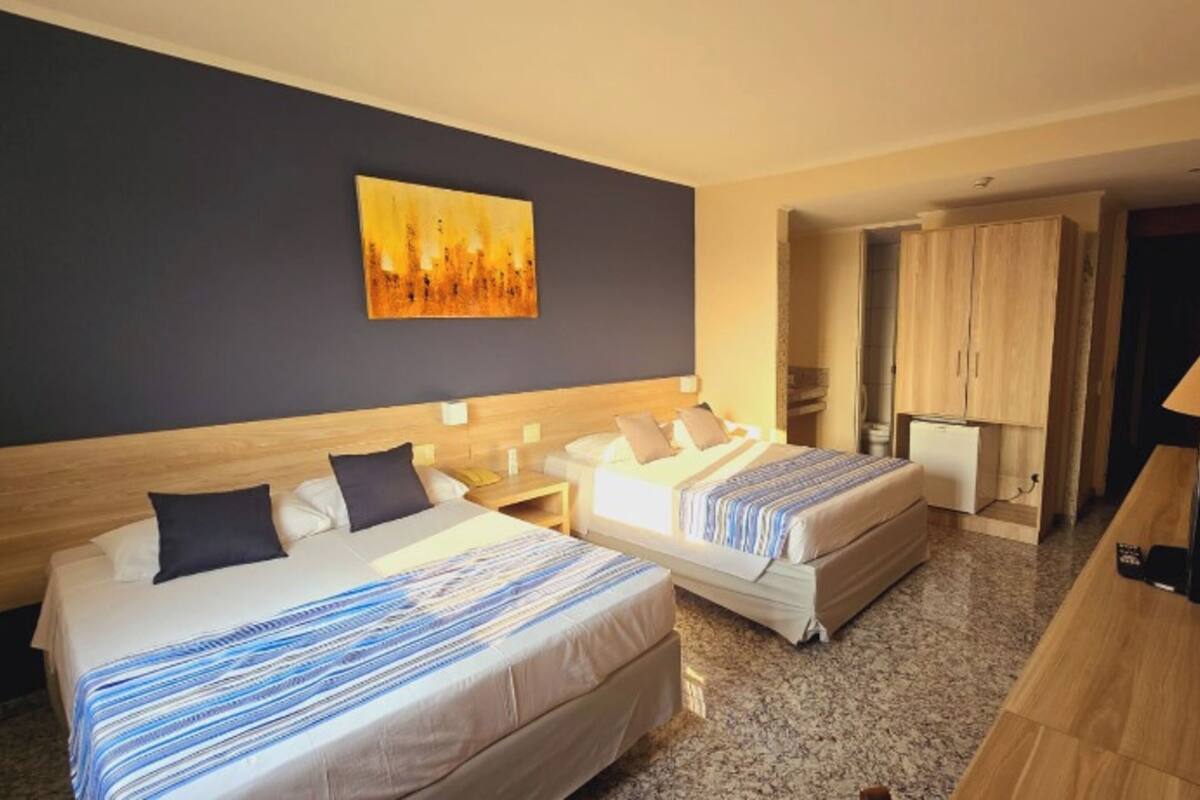 A hotel room with two beds, a painting on the wall, a desk with a lamp, and a wardrobe.
