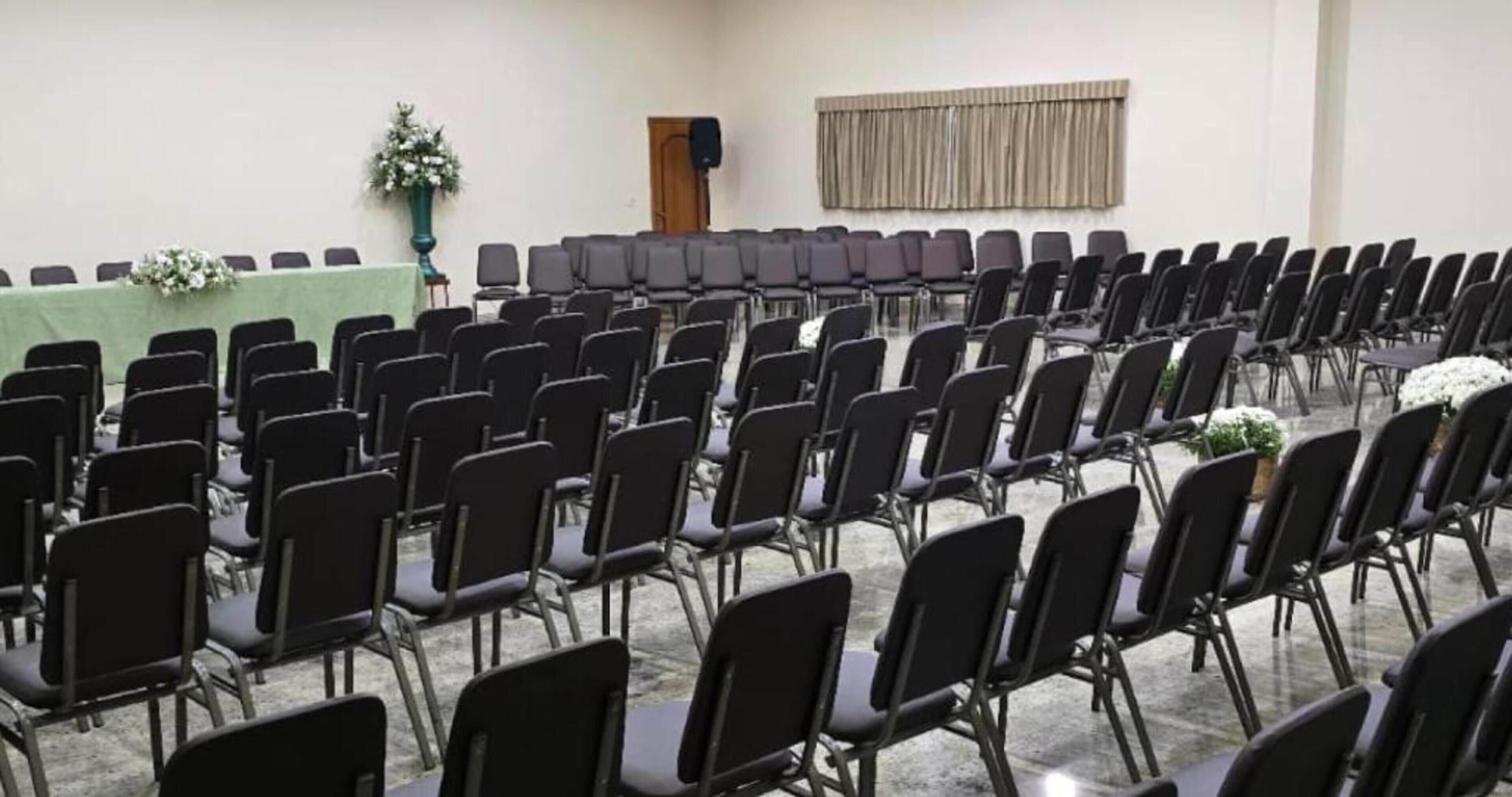Uma sala de conferências com fileiras de cadeiras pretas, uma mesa verde e um alto-falante na parede.