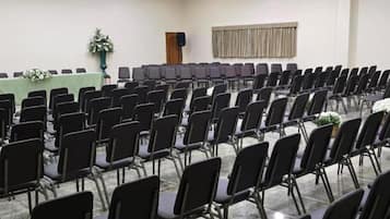 Uma sala de conferências com fileiras de cadeiras pretas, uma mesa verde e um alto-falante na parede.