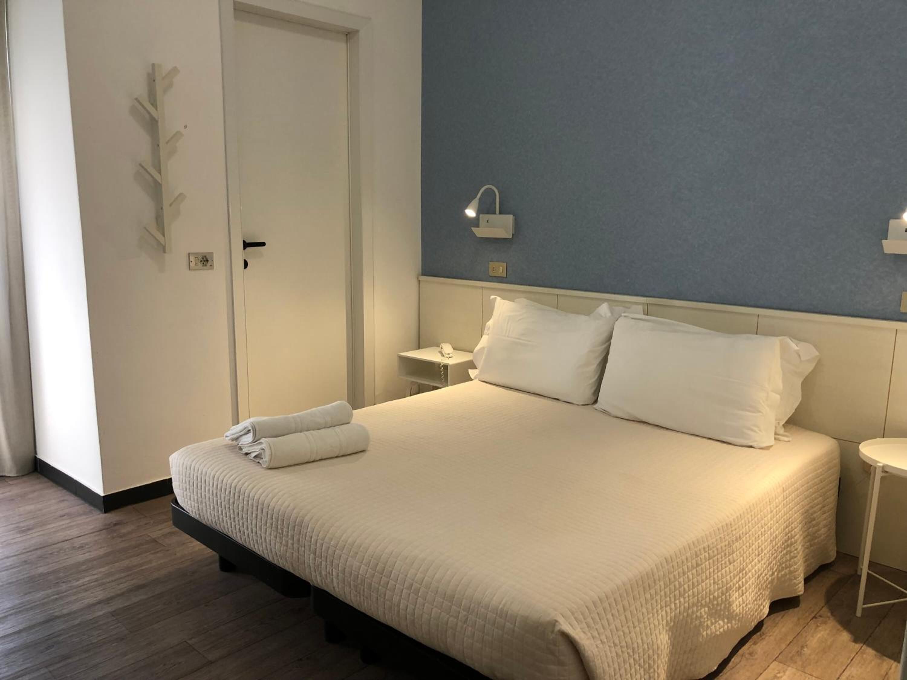 A hotel room with a bed, a bedside table, a nightstand, a wall-mounted phone, and a door.