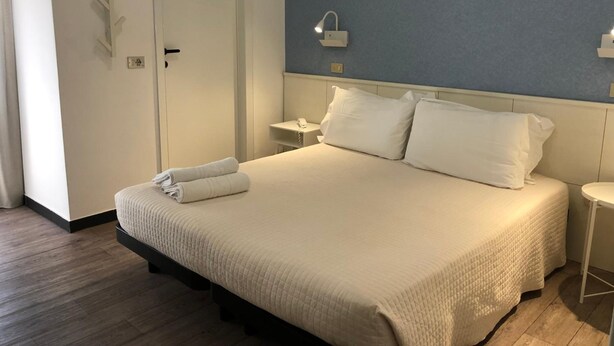 A hotel room with a bed, a bedside table, a nightstand, a wall-mounted phone, and a door.