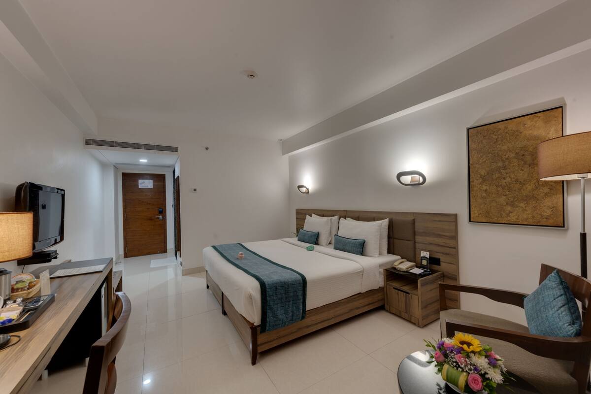 Una habitación de hotel con cama, mesita de noche, lámpara, televisión, escritorio y un arreglo floral.