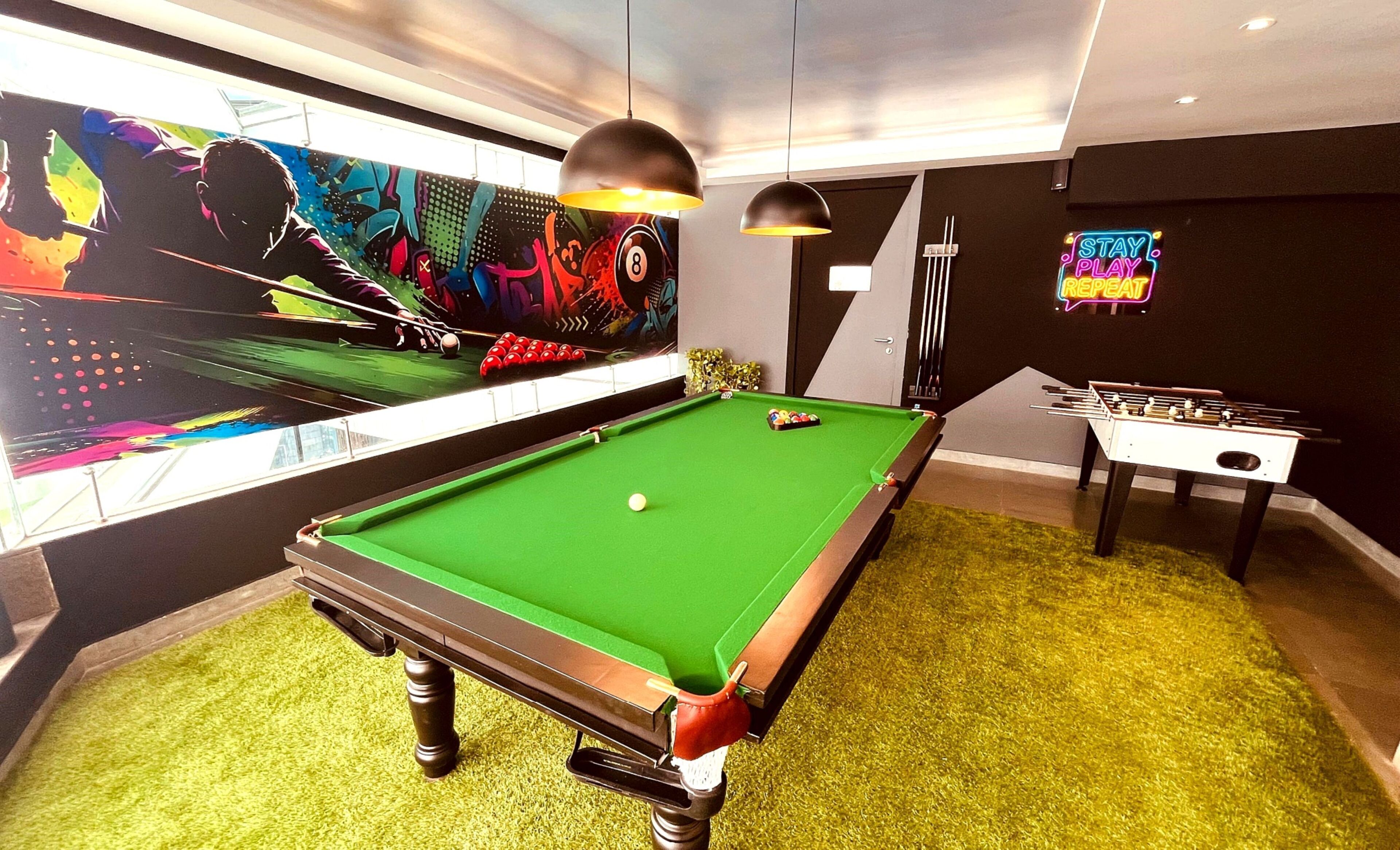 A pool table with a green felt surface, a foosball table, and a colorful mural on the wall.
