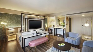 A modern hotel room with a flat-screen TV, a desk with a bench, a sofa, a coffee table, and a small kitchenette. - Hotel Suba International (Mumbai)
