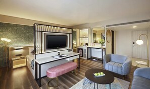 A modern hotel room with a flat-screen TV, a desk with a bench, a sofa, a coffee table, and a small kitchenette. - Hotel Suba International (Mumbai)