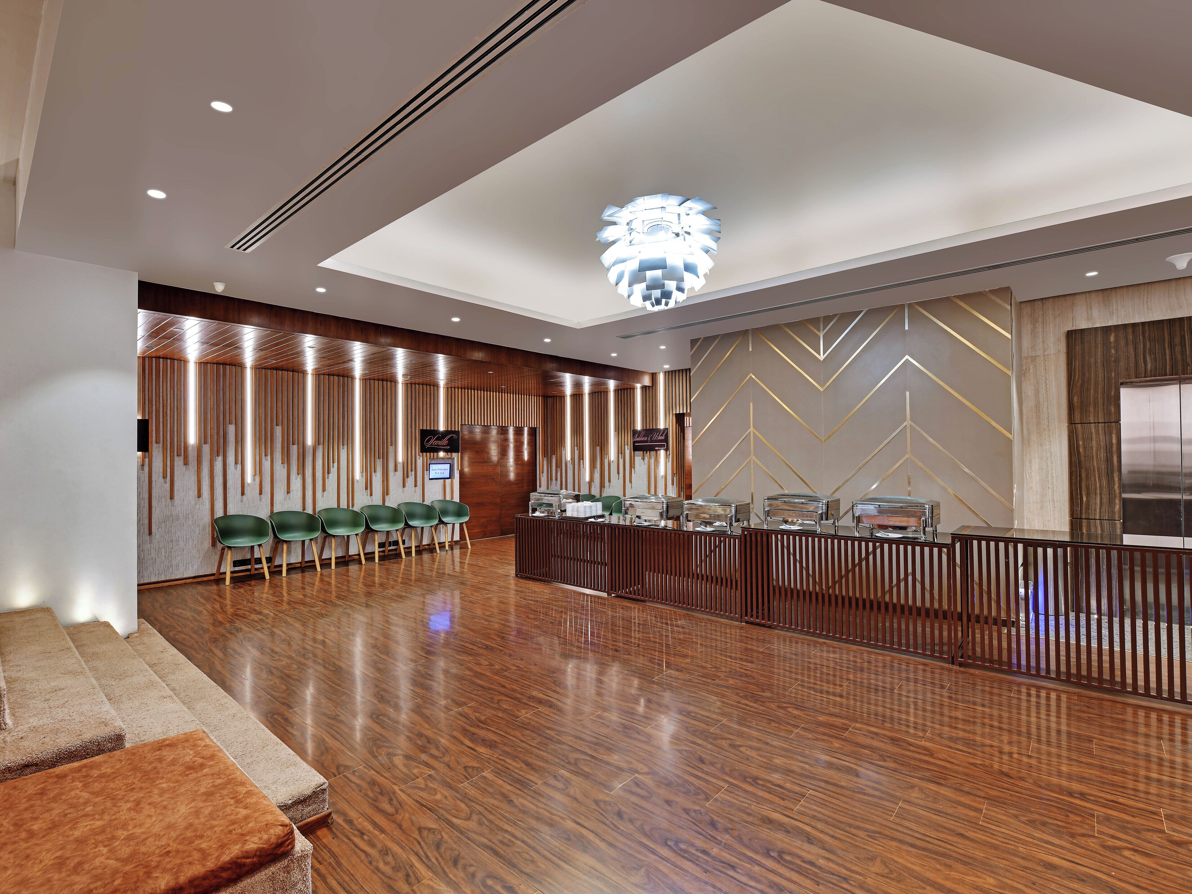 A hotel lobby with a reception desk, green chairs, a wooden floor, and a modern chandelier.