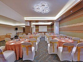 A conference room with round tables, white chairs with yellow sashes, a speaker, and a presentation screen.