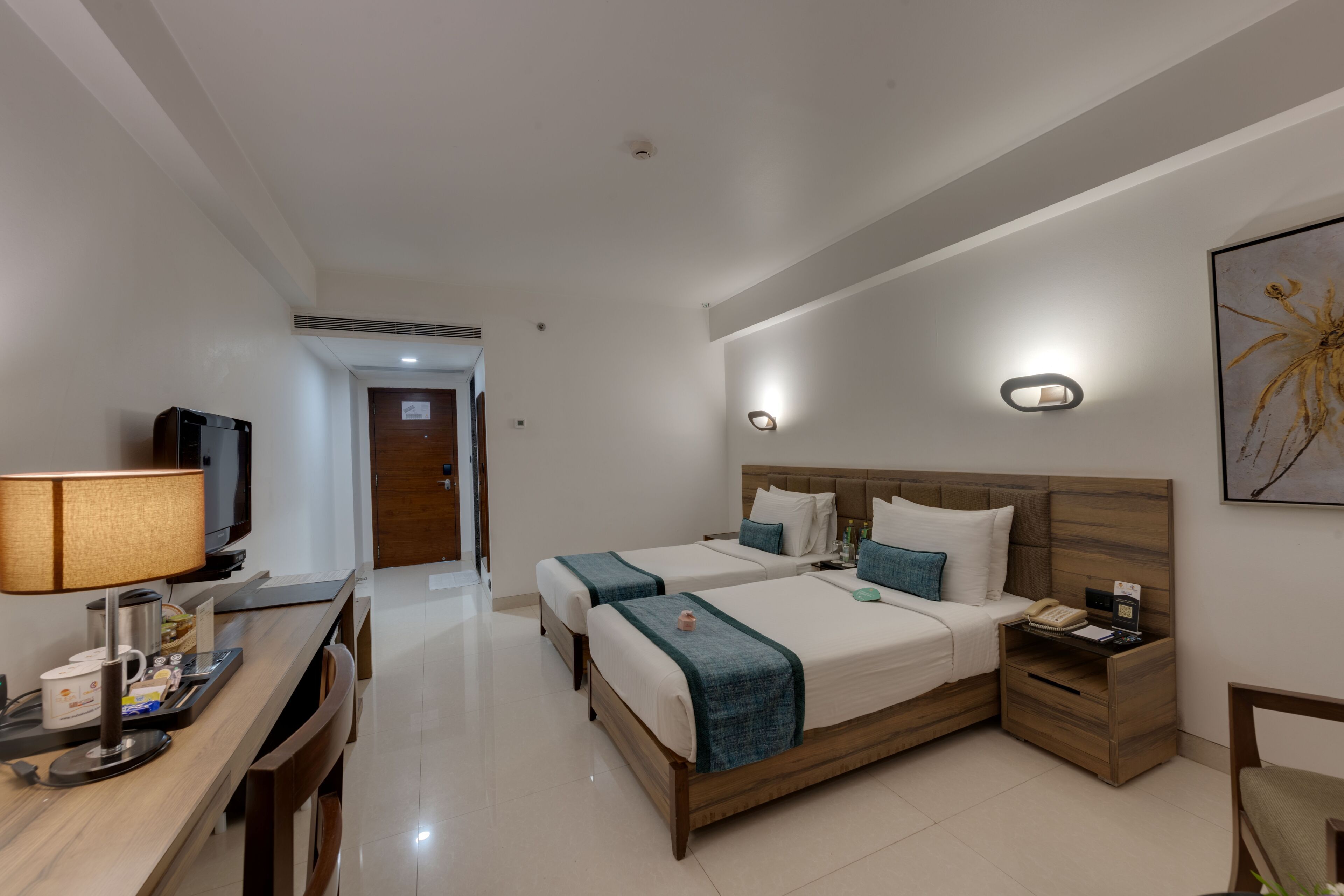 Una habitación de hotel con dos camas, un escritorio con lámpara, televisor y una mesita de noche con teléfono y revista.