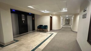 Un pasillo de hotel con piso de mármol, dos ascensores y una zona alfombrada con sillas.