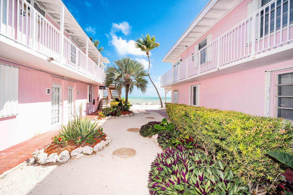 A pathway leading to a beach with pink buildings and palm trees.