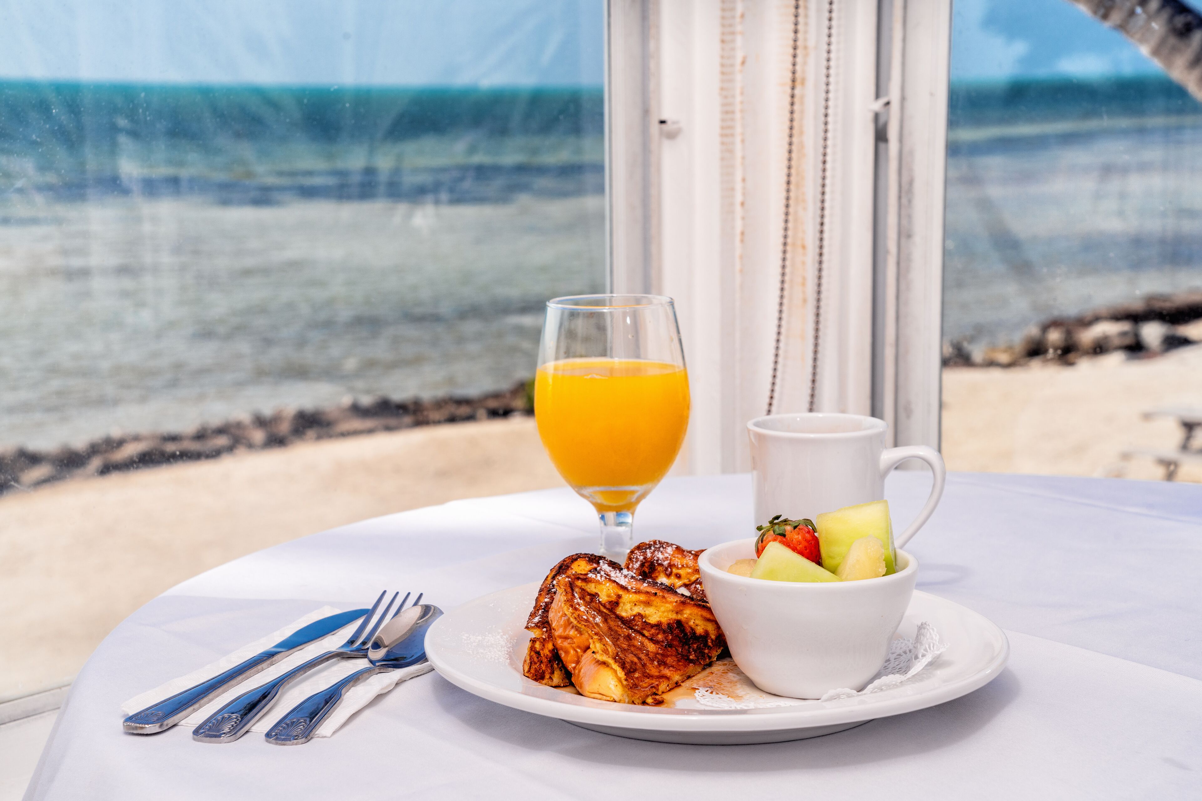 A breakfast setting with toast, fruit, and orange juice on a table by a window overlooking the beach.