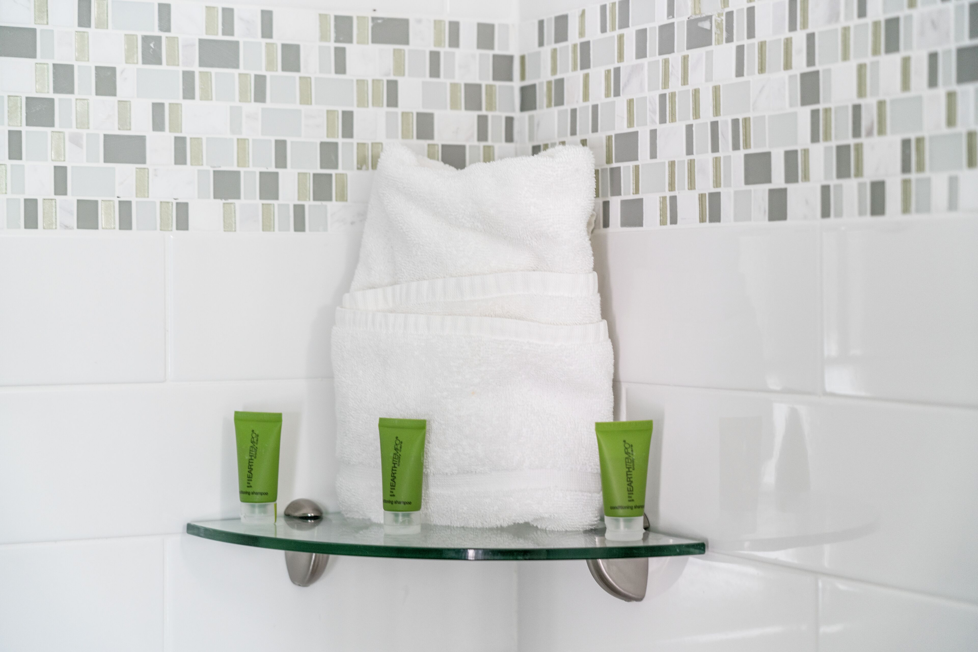 A bathroom with a white towel hanging on a glass shelf, two green tubes of cream, and a mosaic tile wall.