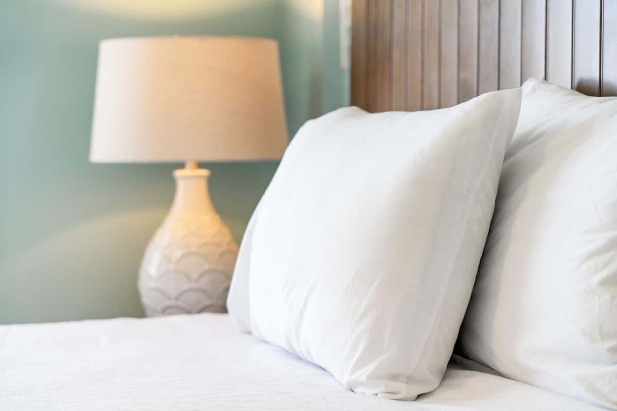 A neatly made bed with white linens and pillows, a white lamp with a scalloped design, and a wooden headboard.