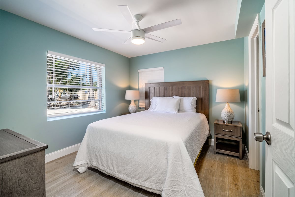A bedroom with a bed, two bedside tables with lamps, a ceiling fan, and a window with blinds.