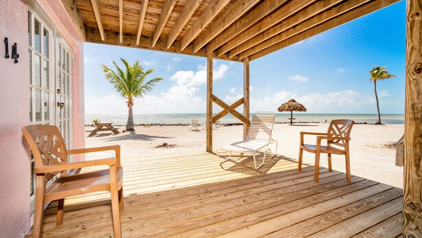 Una terraza de madera con dos sillas y una mesa, con vista a una playa con palmeras y el océano.