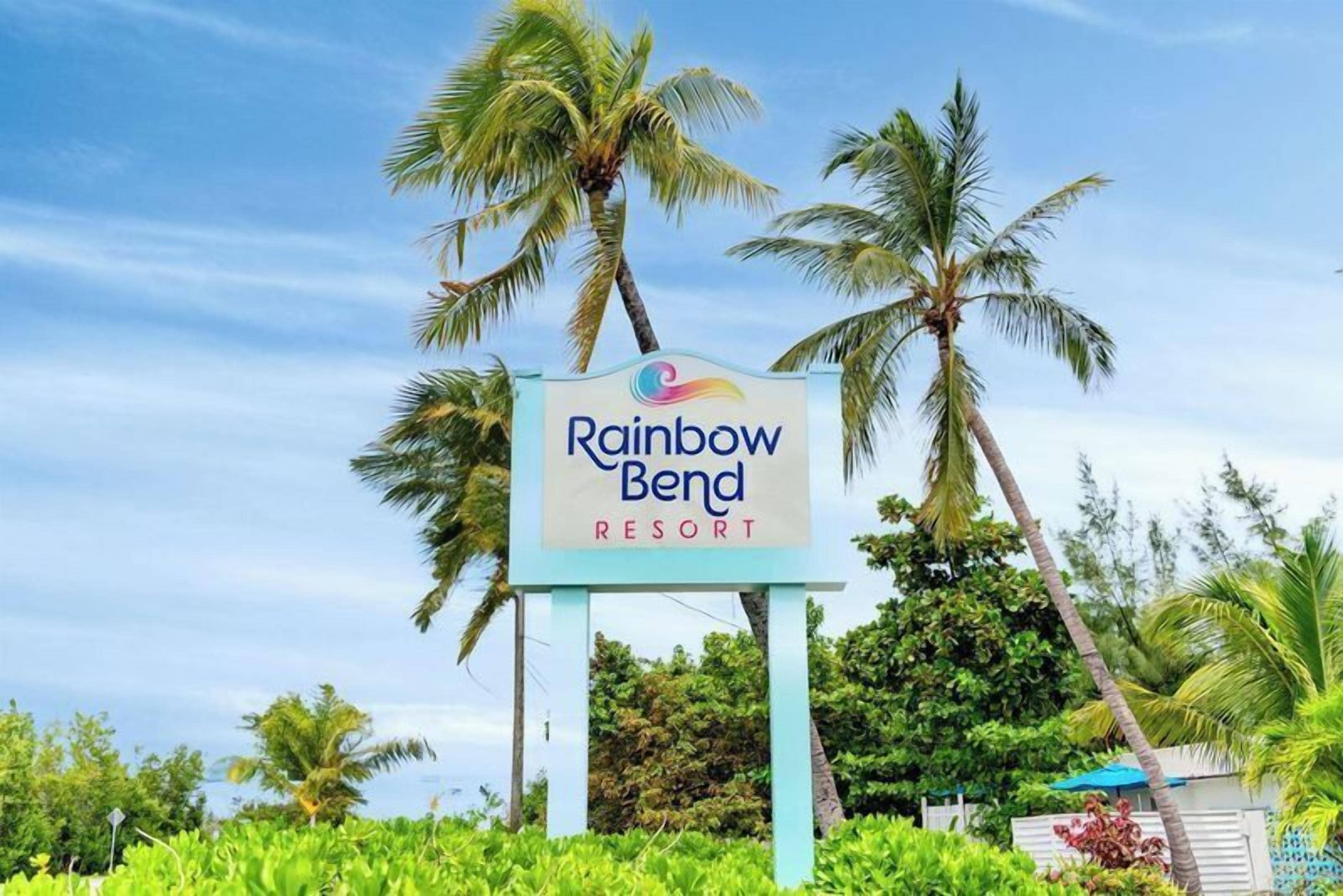 A sign for Rainbow Bend Resort surrounded by palm trees and greenery.