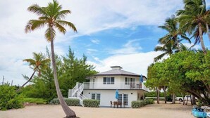 Una casa frente a la playa de dos pisos con balcón, rodeada de palmeras y una playa de arena.