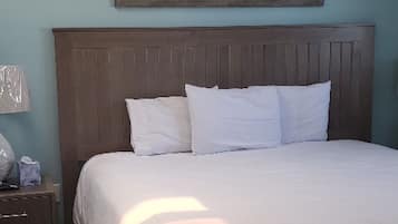 Un lit bien fait dans une chambre avec une tête de lit en bois, un tableau représentant une scène de plage au-dessus, et une table de chevet en bois avec une lampe et une boîte à mouchoirs.