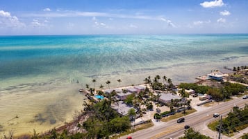An aerial view of a coastal area with clear turquoise waters, a road with vehicles, and a cluster of buildings with a swimming pool.