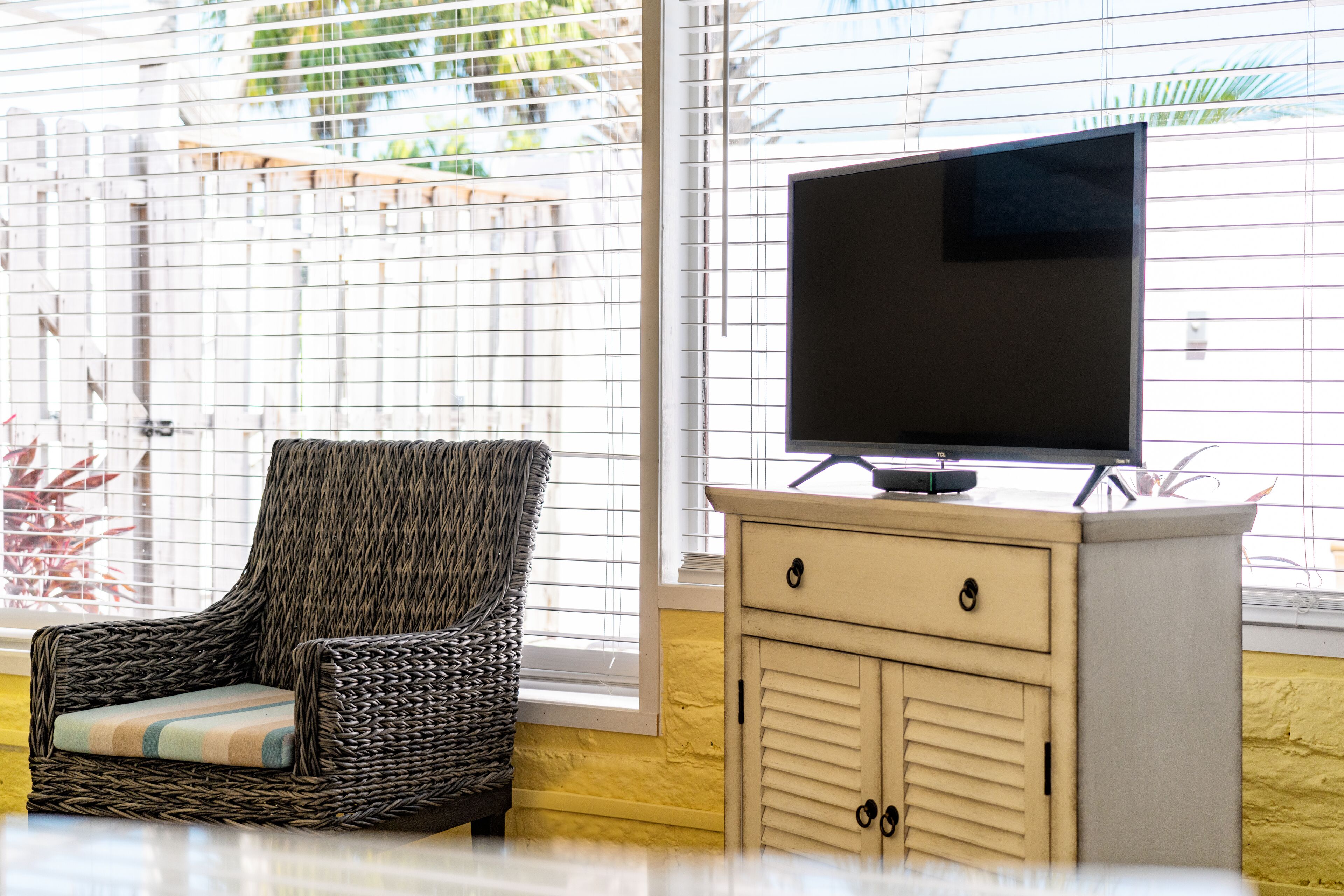 A room with a wicker chair, a television on a cabinet, and a window with blinds.