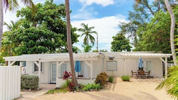 Un edificio blanco de un piso con un patio cubierto, rodeado de vegetación tropical y palmeras.