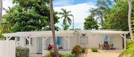 Un edificio blanco de un piso con un patio cubierto, rodeado de vegetación tropical y palmeras.