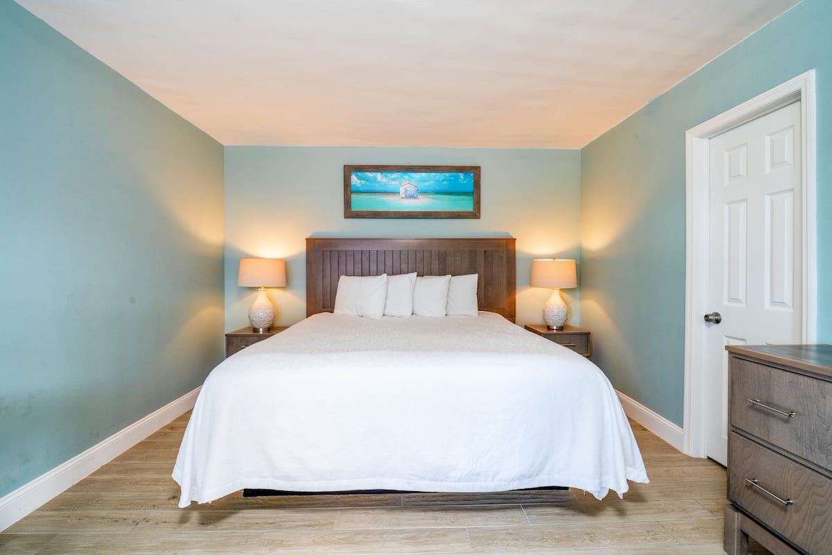 A neatly made bed in a room with light blue walls, a wooden headboard, and a framed picture above the bed. There are two bedside tables with lamps.