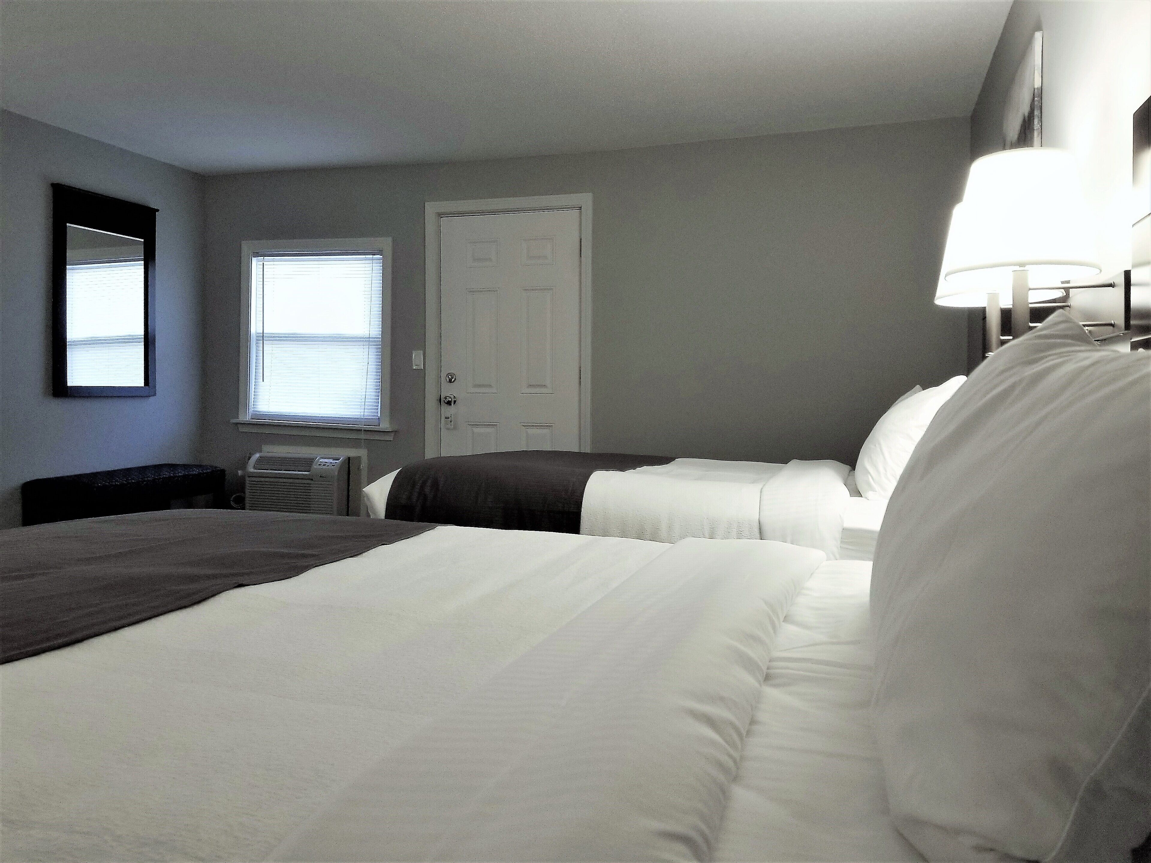 A hotel room with two beds, a window, a door, and a wall-mounted lamp.