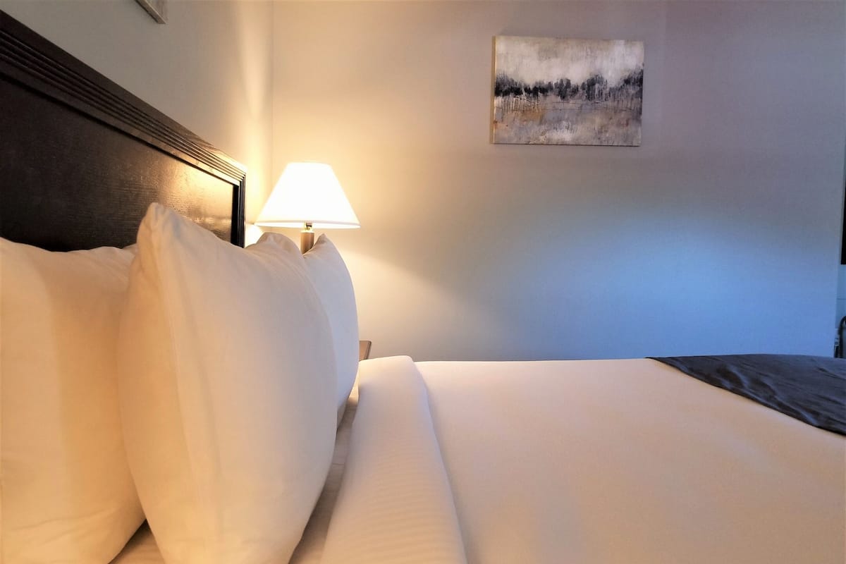 Ein ordentlich bezogenes Bett mit weißen Laken, eine Lampe daneben und ein Gemälde an der Wand.