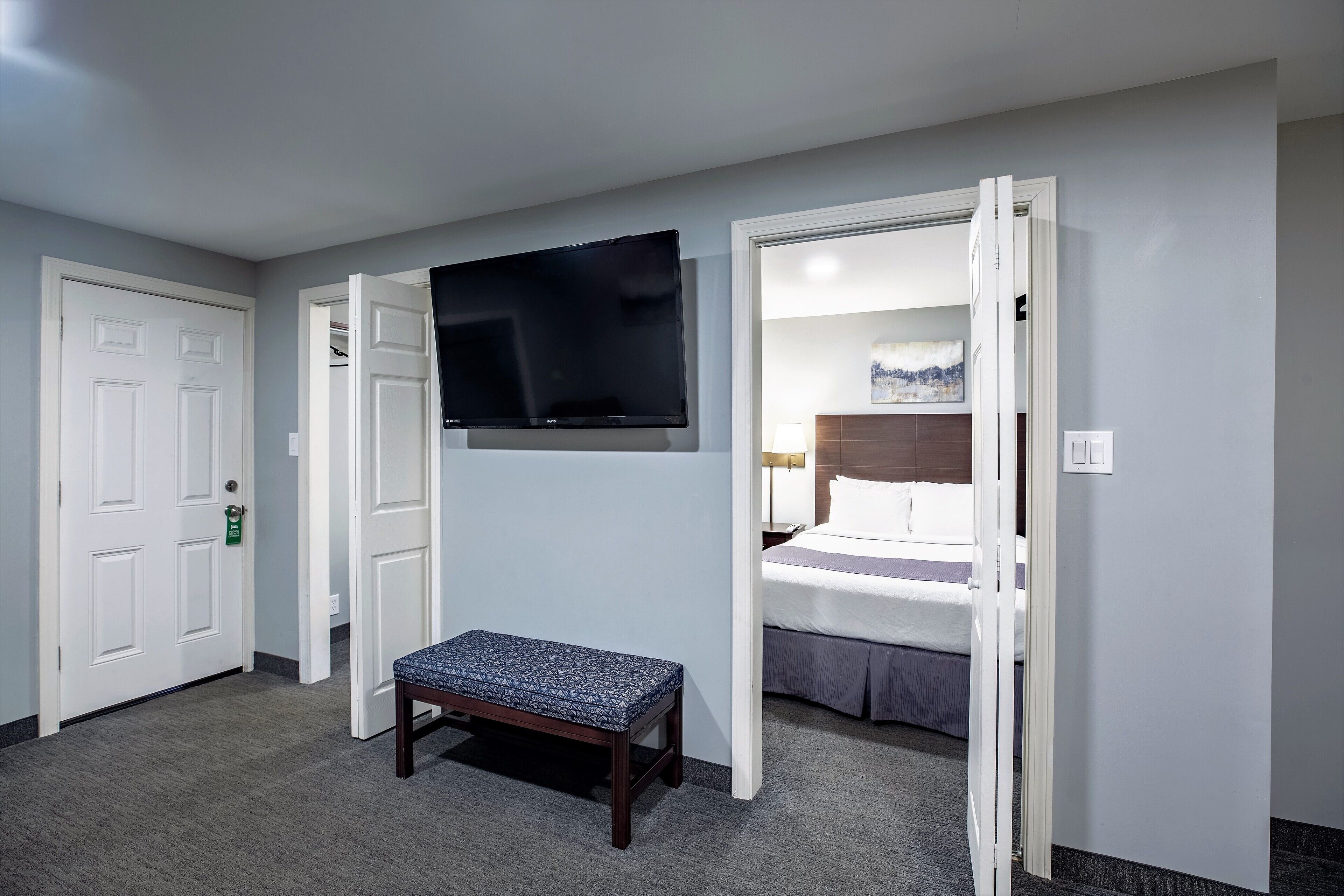 A hotel room with a bed, a television mounted on the wall, a bench, and a door leading to another room.
