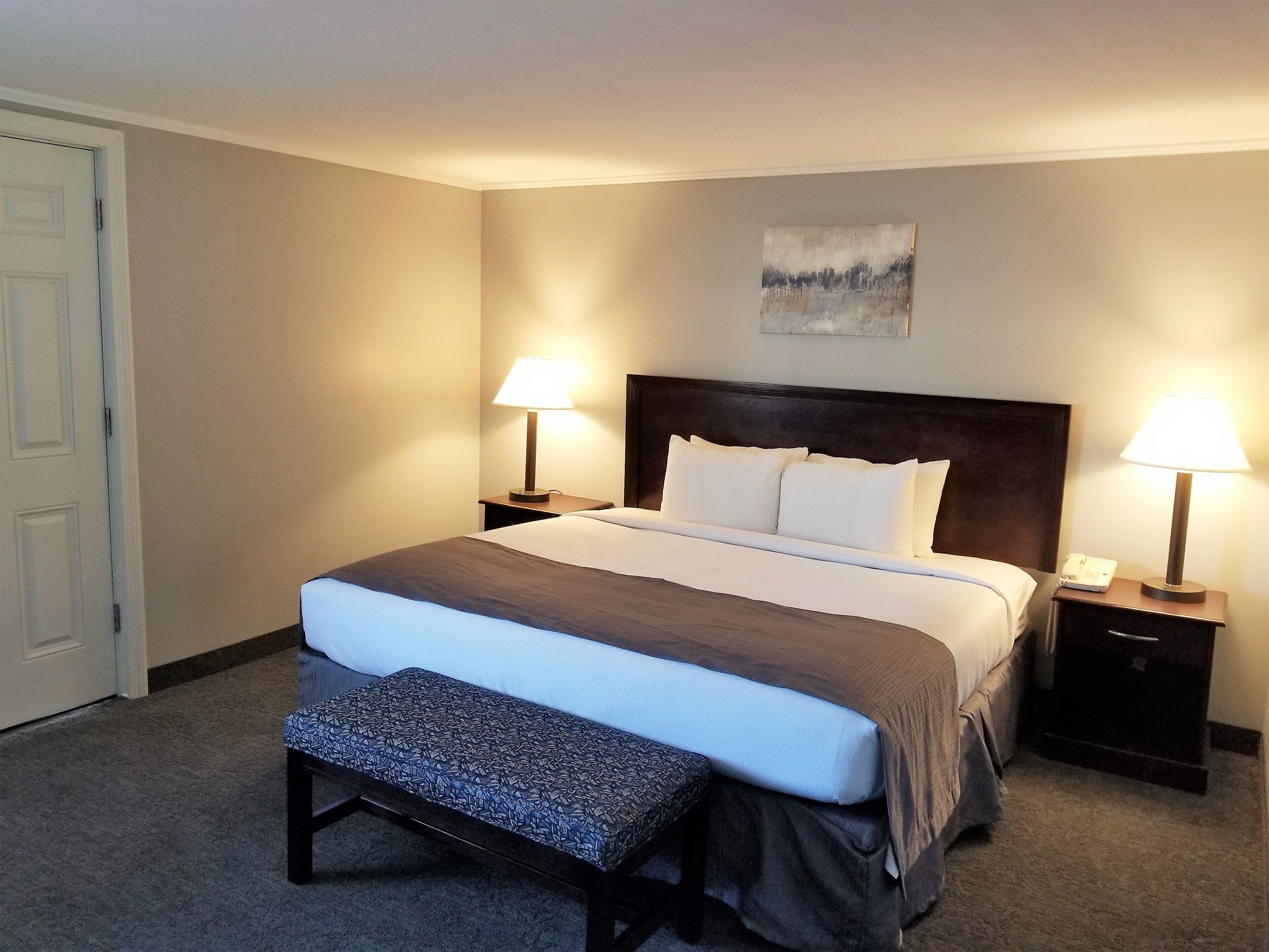 A neatly made bed with a headboard, two bedside lamps, a bench, and a painting on the wall.
