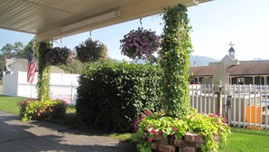 A covered outdoor area with hanging flower baskets, a brick planter with flowers, and a white picket fence. - Mt Madison Inn & Suites (Gorham)