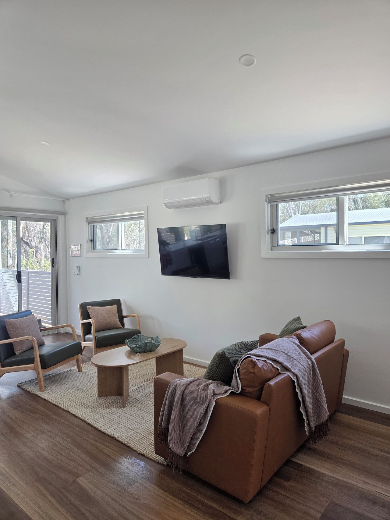 A modern living room with a flat-screen TV mounted on the wall, a brown leather sofa, two armchairs, a wooden coffee table, and a large window with a view of a house.