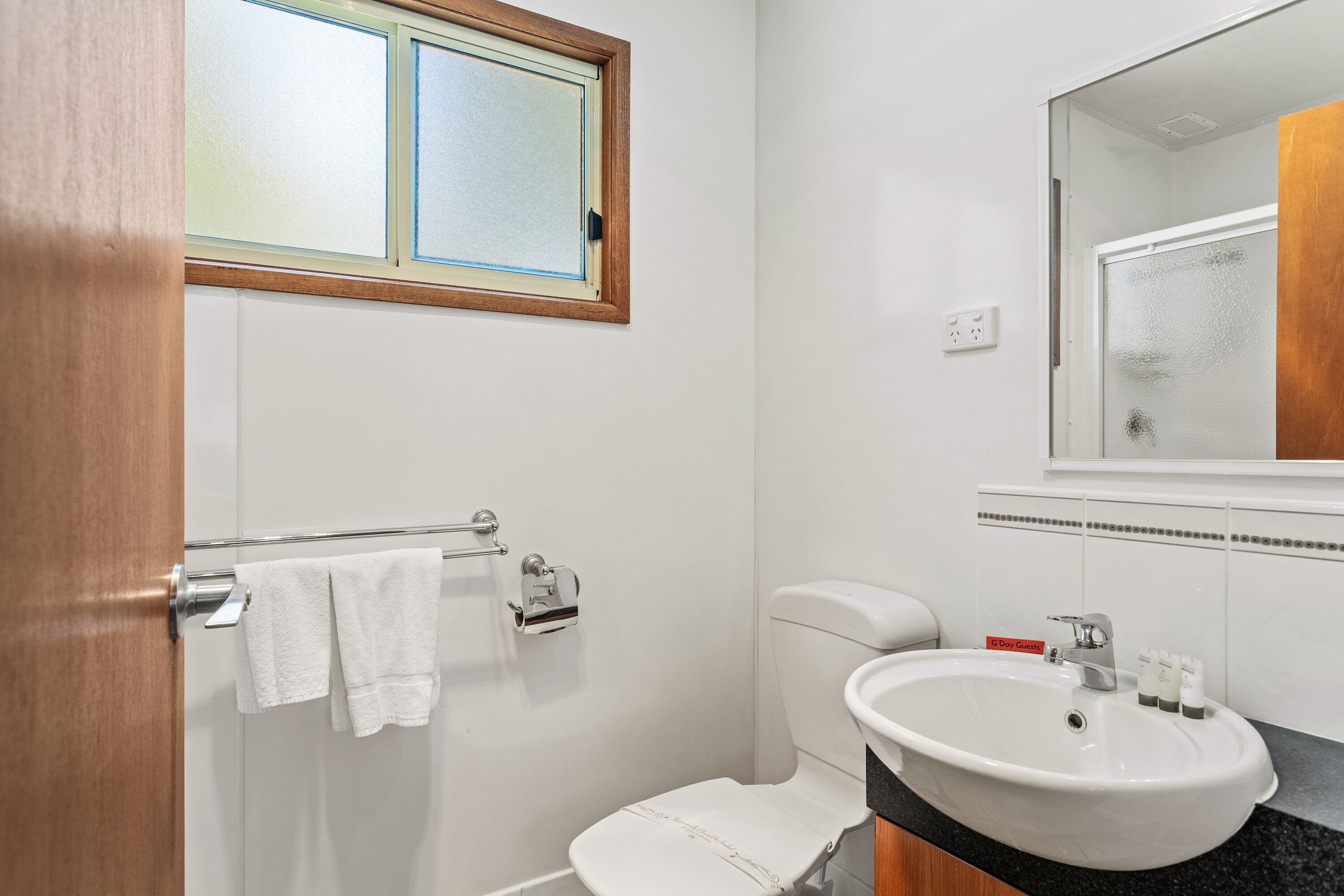 Un baño con inodoro, lavamanos, perchero para toallas y una ventana sobre el inodoro.