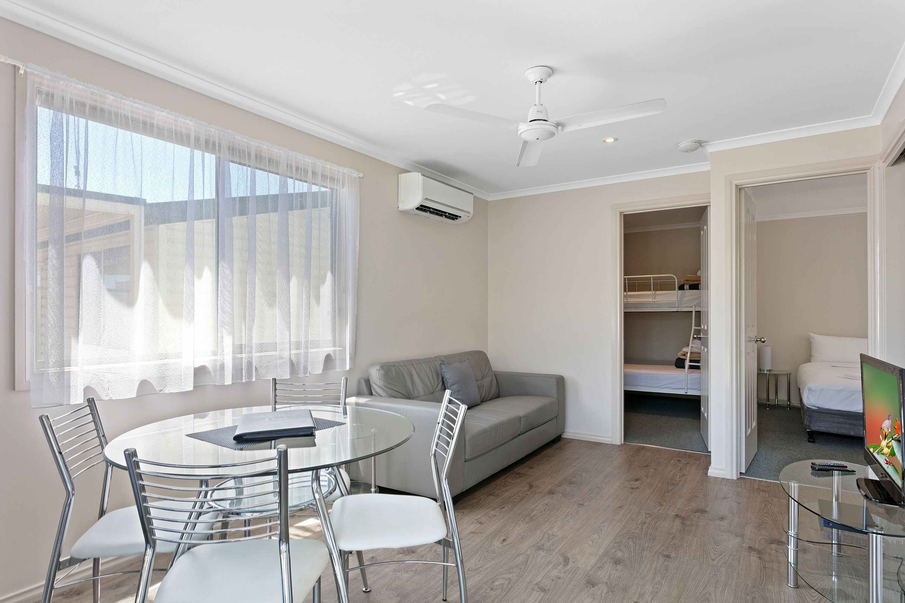A modern room with a round glass table, white chairs, a grey sofa, a ceiling fan, and an air conditioning unit.