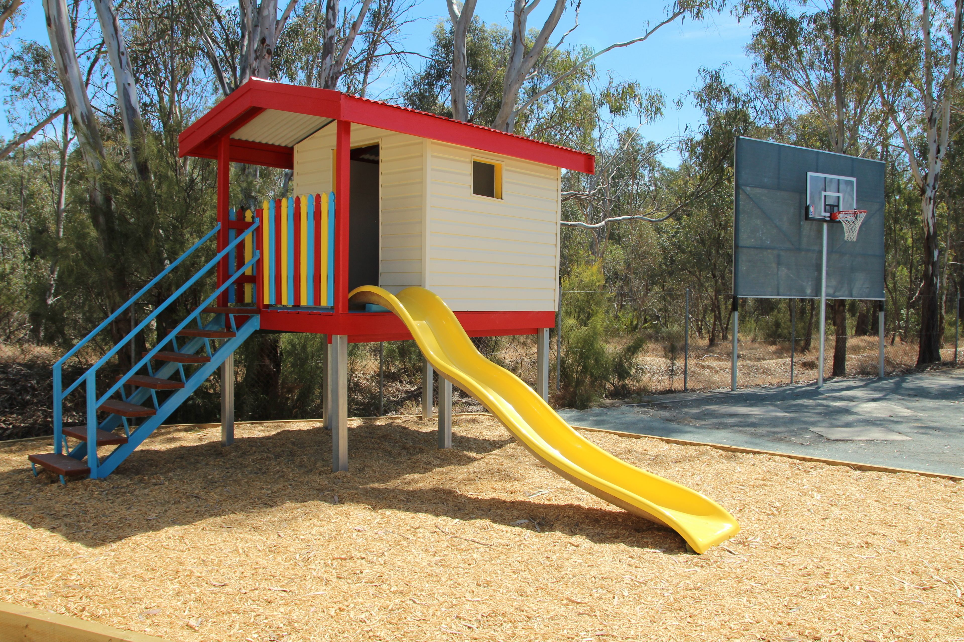A playground with a yellow slide, a red and yellow playhouse, and a basketball hoop.