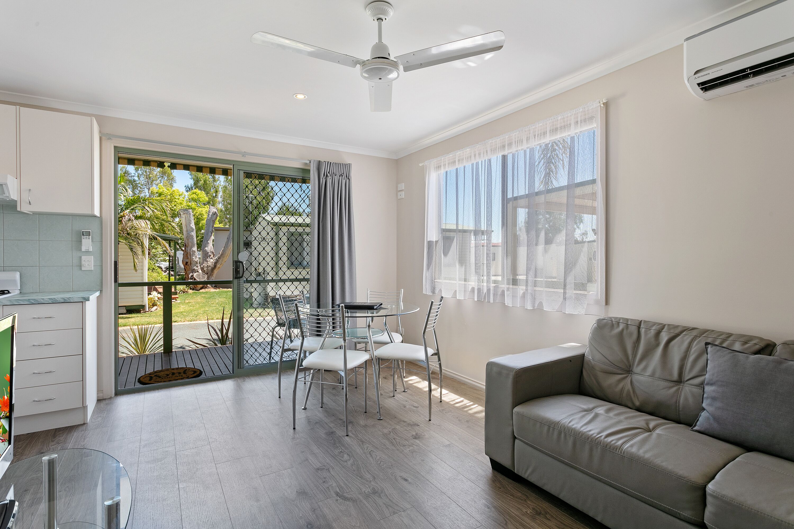 A modern living room with a grey sofa, a dining table with chairs, a ceiling fan, and a sliding glass door leading to a balcony.