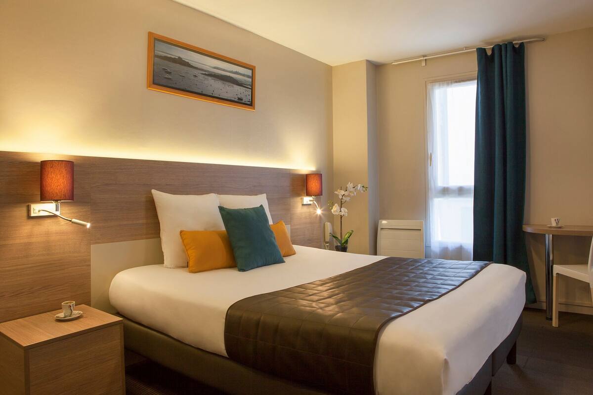 Ein Hotelzimmer mit einem großen Bett, Nachttischlampen, einem kleinen Tisch mit einer Tasse und einem Fenster mit Vorhängen.