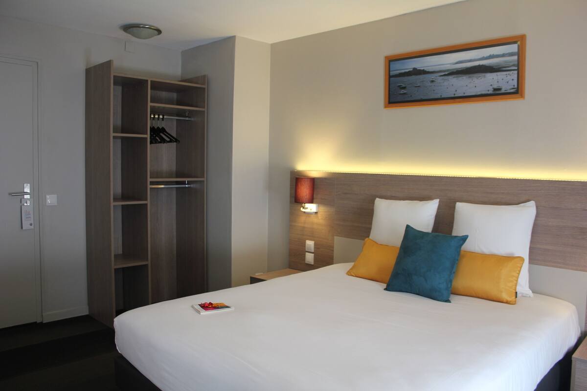 Ein Hotelzimmer mit Bett, Nachttisch, Wandlampe, einem gerahmten Bild und einem Kleiderschrank.