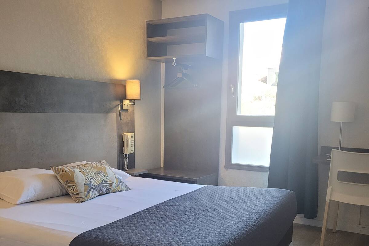 Ein Hotelzimmer mit einem großen Bett, einem Nachttisch mit Lampe, einem Kleiderhaken, einem Sessel und einem Fenster mit Vorhängen.
