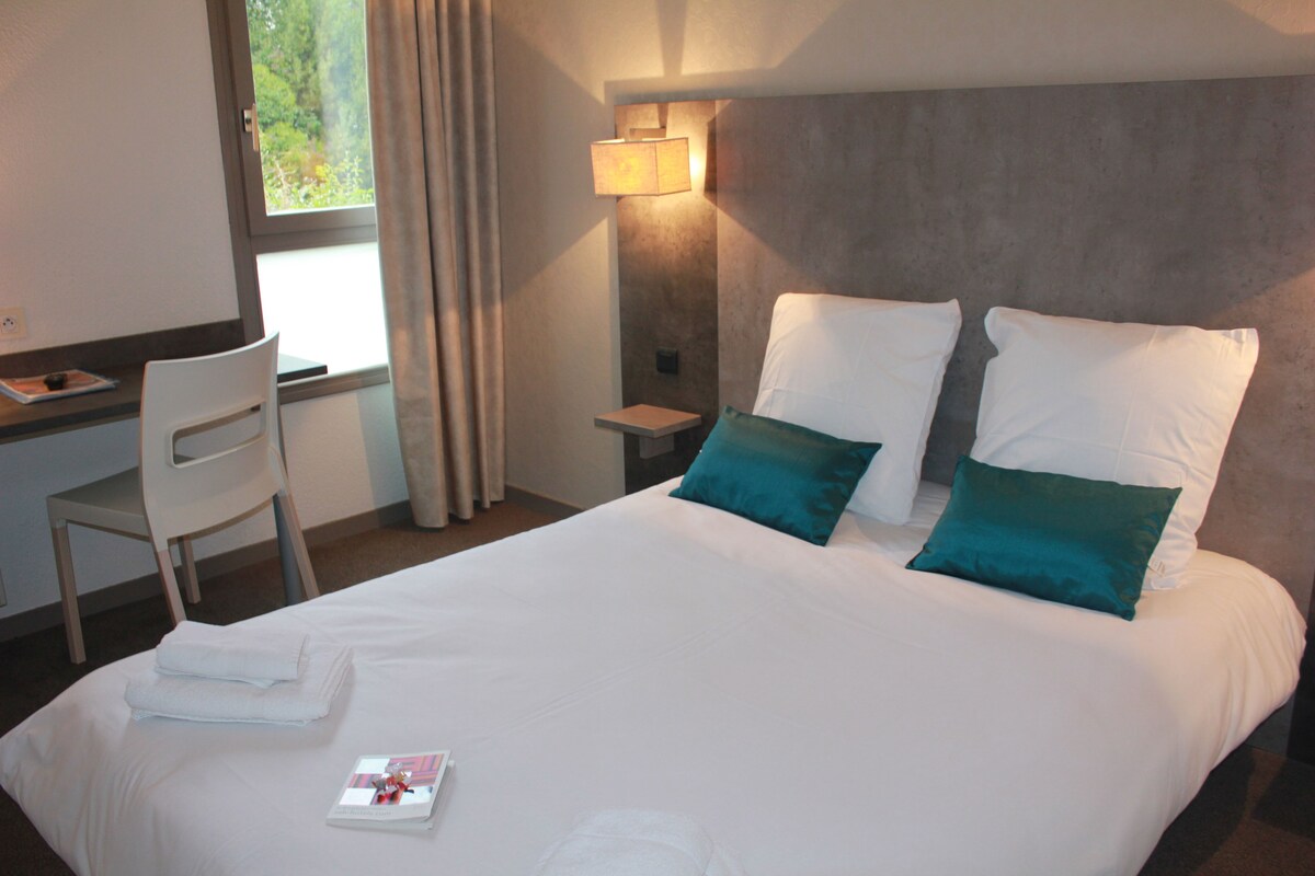 Una cama bien hecha con dos almohadas, una mesita de noche con una lámpara, una silla y una ventana con cortinas.